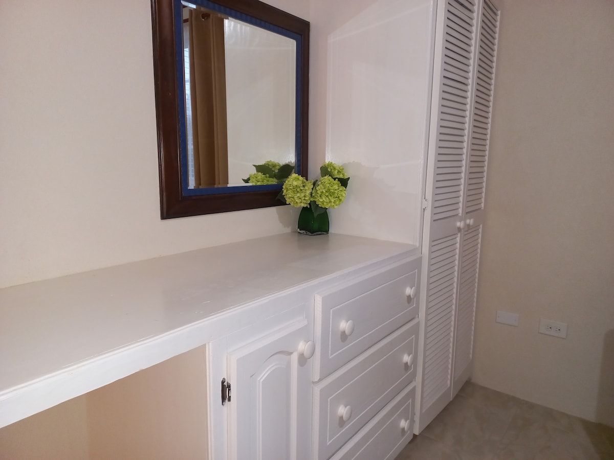 A simple white dressing table is positioned against a light wall, complemented by a framed mirror above it. A small arrangement of green flowers adds a touch of color. Nearby, a white cabinet incorporates multiple closed storage spaces.