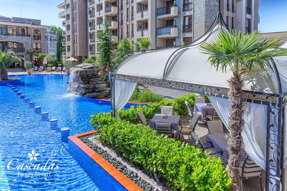 A tranquil pool area features a spacious blue water pool surrounded by lush greenery. A shaded cabana with tables and chairs provides a cozy space for relaxation, while stone landscaping enhances the resort's serene atmosphere.