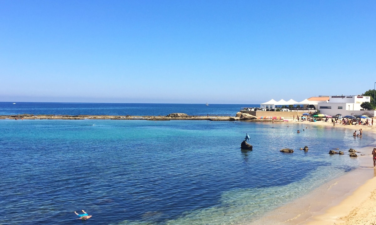 The image captures a serene beach scene with crystal-clear waters gently lapping against the shore. Sunbathers can be seen enjoying the sun, while a few small boats are anchored nearby. A vibrant blue sky stretches overhead, enhancing the coastal ambiance.