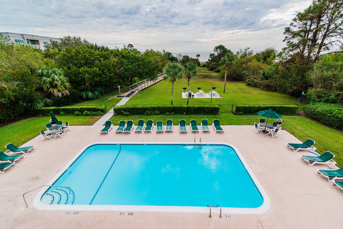 An outdoor swimming pool is surrounded by green lounge chairs, arranged neatly along the pool's edge. Lush landscaping includes palm trees and well-maintained grass areas, offering a serene environment. A path leads from the pool area to additional outdoor seating in the background.