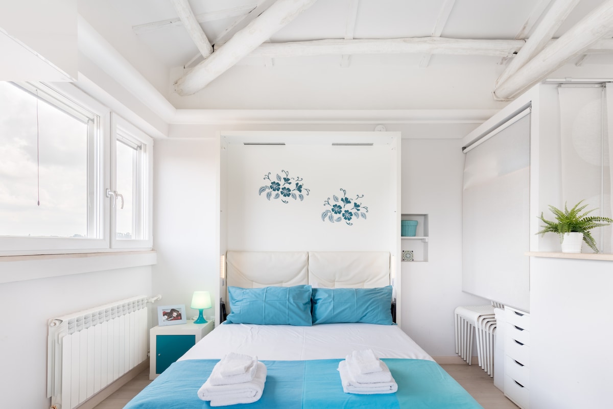 A bright bedroom is highlighted by a spacious bed with blue and white linens. Soft blue accents and floral wall art add a touch of color. Natural light floods the room through large windows, and a small lamp provides gentle illumination. The modern design features exposed wooden beams overhead.