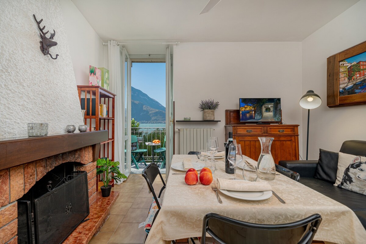 A cozy living area features a fireplace and a dining table set for four. A contemporary black sofa is positioned nearby, with a television displaying scenic views. Large glass doors provide access to a balcony, offering views of the surrounding mountains.