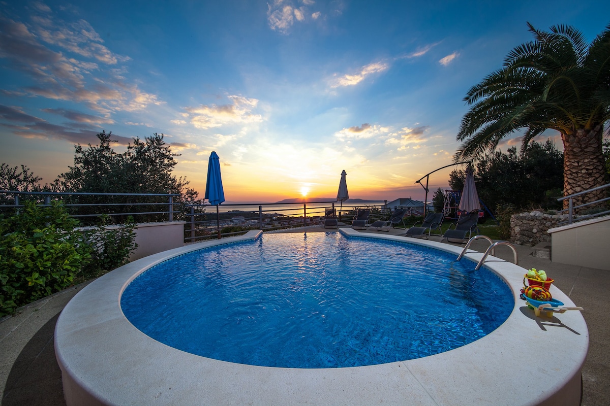 A private pool is situated at the villa, surrounded by lush greenery and several sun loungers with umbrellas. The sunset creates a calming atmosphere over the water, with the sea visible in the background.