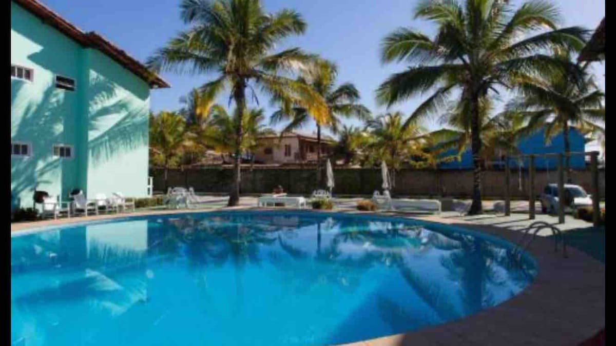 A clear swimming pool is surrounded by palm trees and lounge chairs. The vibrant blue water reflects the bright sky and greenery. Nearby, shaded areas feature tables and umbrellas, offering a relaxed atmosphere for guests.