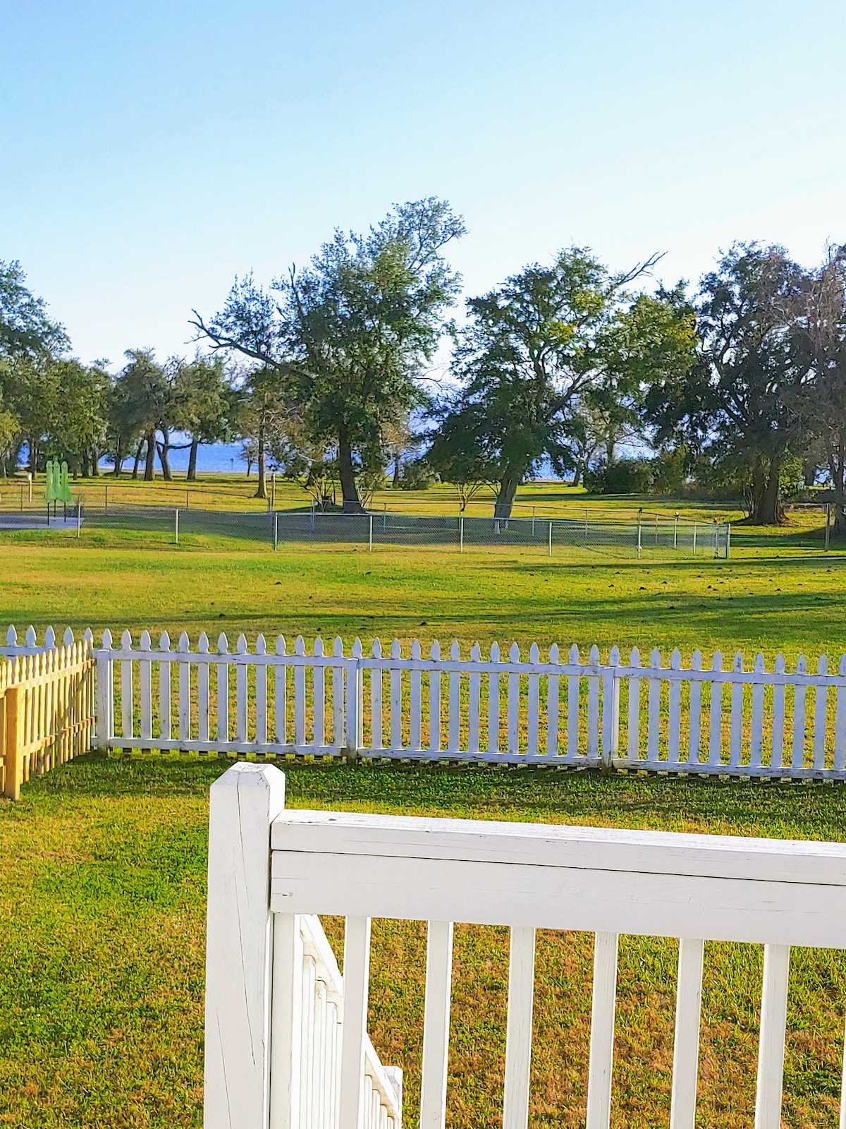 A view of the spacious green lawn stretches beyond a white picket fence, leading to trees and a tranquil body of water in the distance. The scene is brightened by sunlight and clear skies, offering a serene atmosphere.