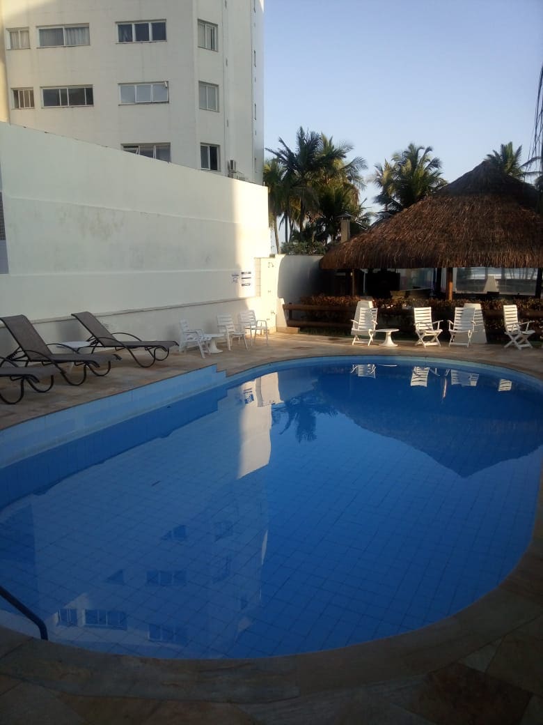 A rectangular swimming pool is surrounded by lounge chairs, providing a space for relaxation. A thatched-roof gazebo is visible, offering shade nearby. Palm trees frame the area, and sunlight reflects off the calm water, creating an inviting atmosphere for leisure.