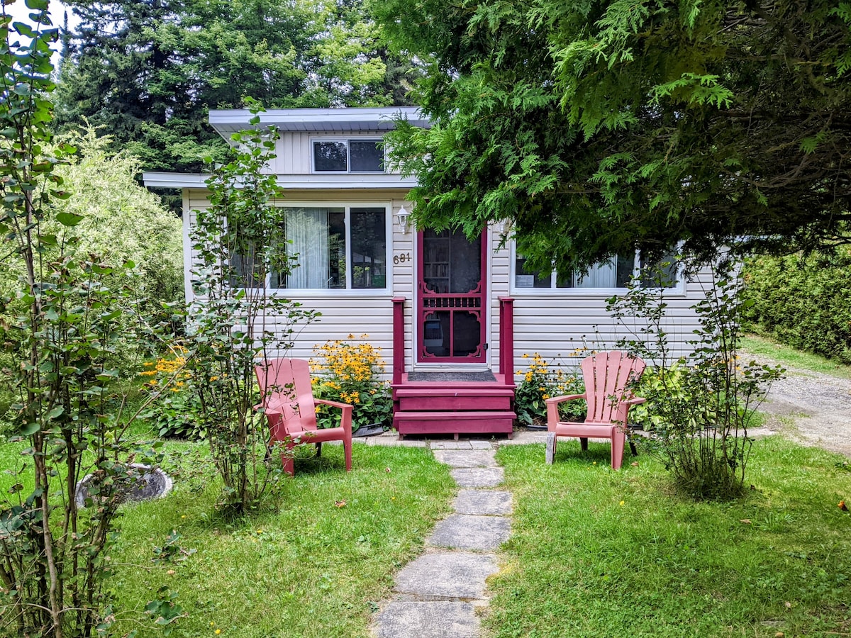 A charming chalet is featured, surrounded by greenery and flower beds. A pathway leads to the entrance, flanked by two red Adirondack chairs. Large windows provide views of the outdoor landscape, and the front porch is accessible via a few steps.