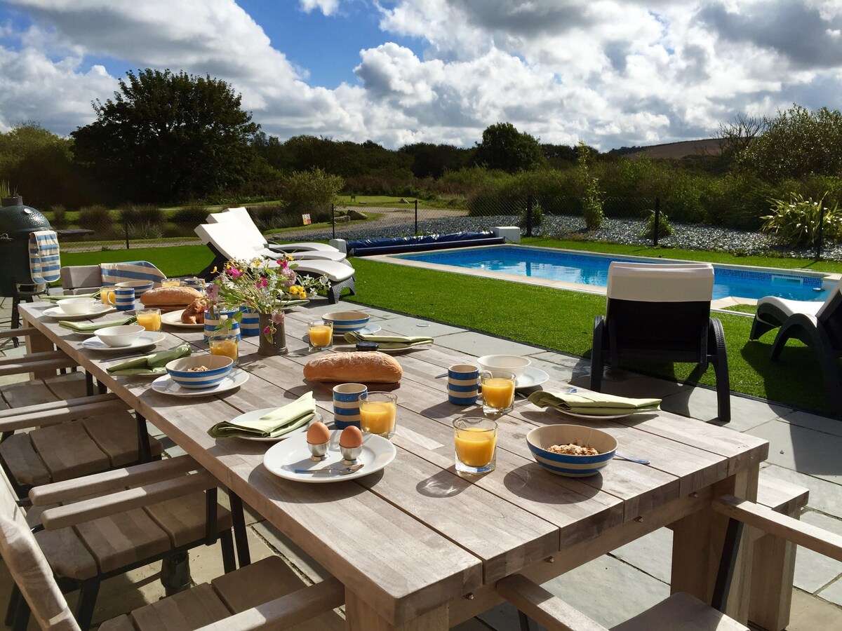 An outdoor dining table is set for breakfast, featuring bowls, glasses of orange juice, and various bread items. The private heated pool is visible in the background, surrounded by grass and lush greenery under a partly cloudy sky.