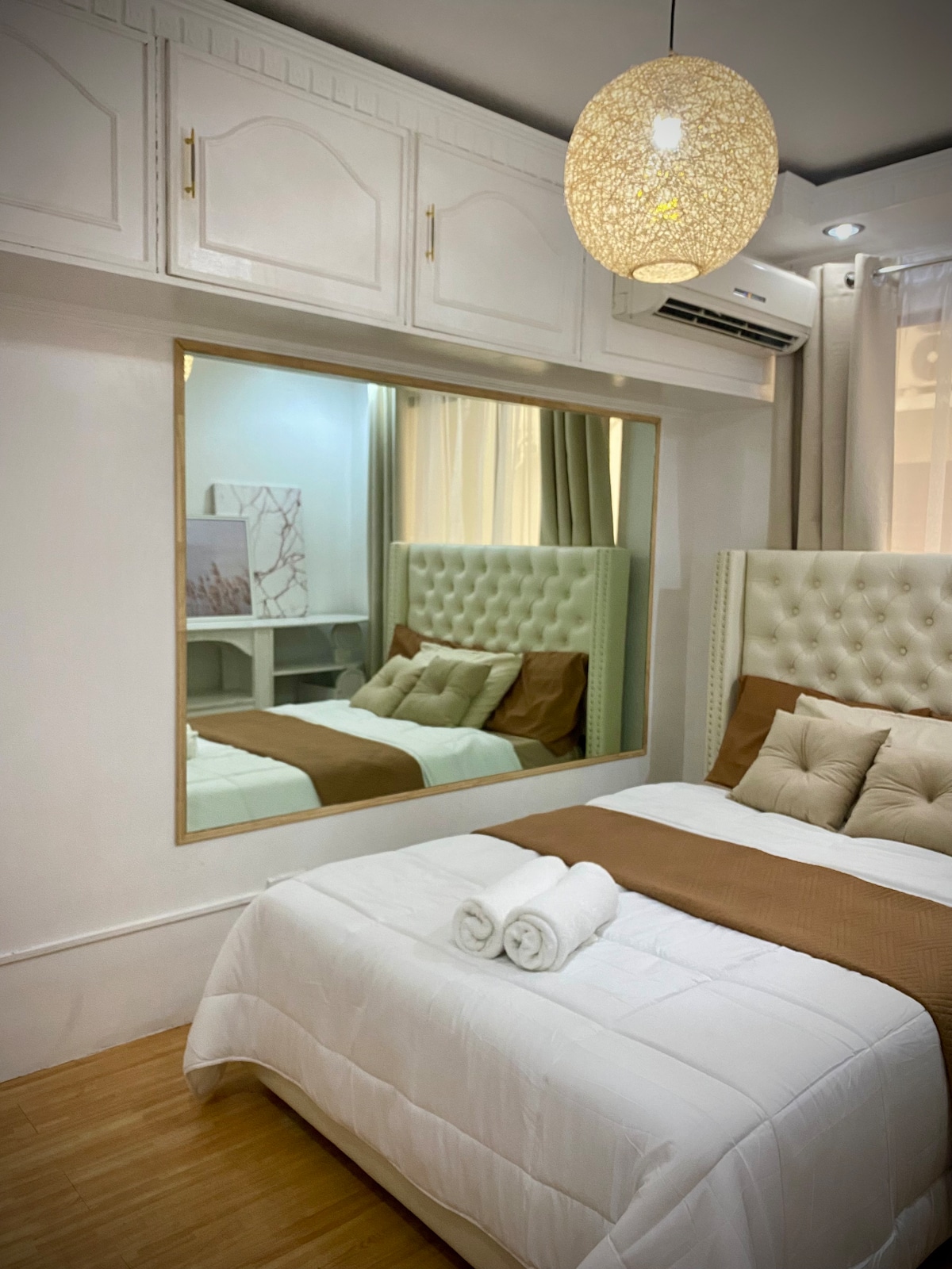 A bright sleeping area features a spacious bed with a tufted headboard and neutral-toned bedding. A mirror is positioned above a light wood dresser, reflecting the soft lighting of the room. Decorative pillows add a touch of comfort, complemented by a pendant light above.