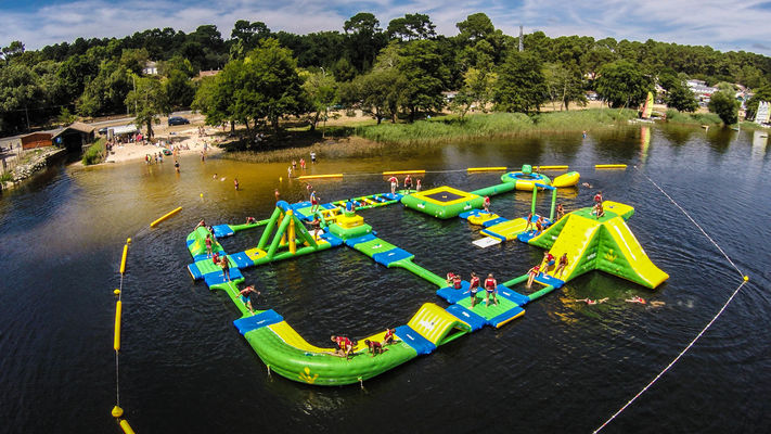 An expansive floating water park is set on a calm lake, featuring colorful inflatable structures in green and yellow. Visitors can be seen enjoying various attractions, including slides and obstacle courses, with people swimming nearby. Lush trees frame the serene backdrop.
