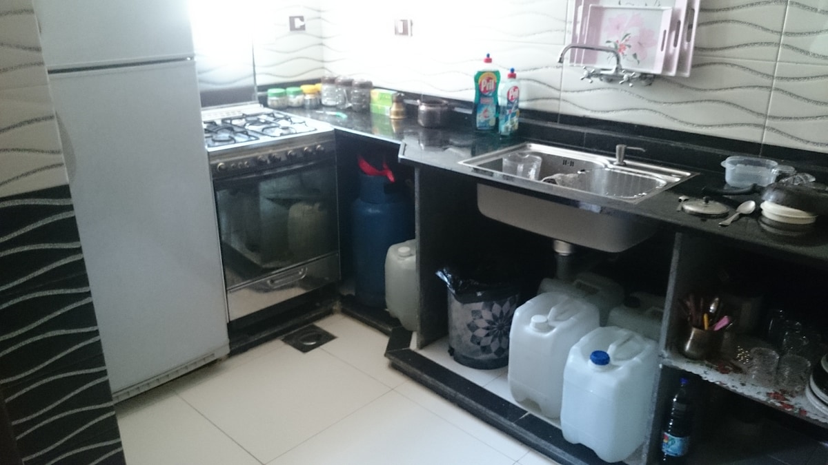 The kitchen features a gas stove and a double sink, with storage cabinets and a countertop displaying various cleaning supplies. Several containers of water are arranged neatly on the floor, and a refrigerator stands against one wall.