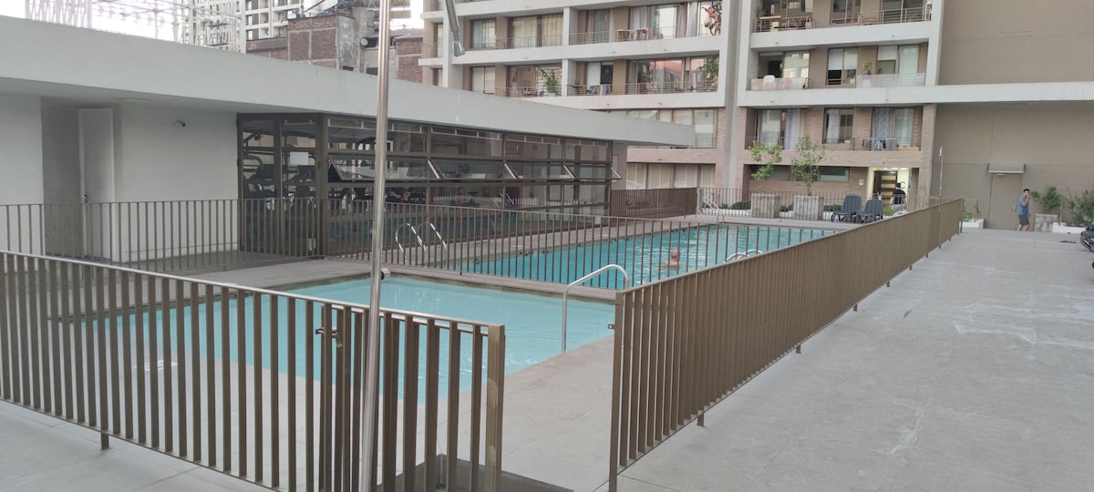 A communal swimming pool is framed by a modern deck and a safety railing. The pool area features sun loungers alongside the water, with the surrounding buildings providing a backdrop. Clear blue water reflects the light, creating a calm ambiance.