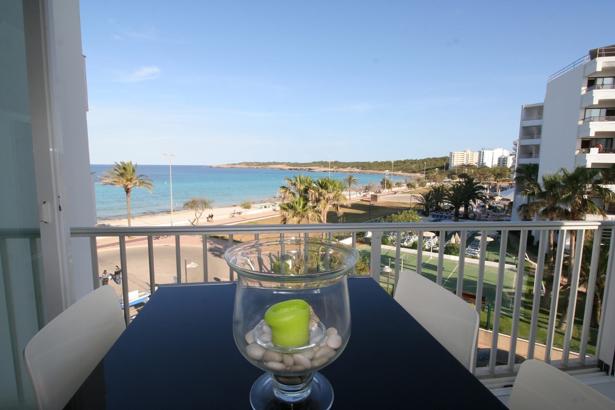 An open terrace reveals a stunning view of the beach and sea, framed by palm trees and coastal greenery. A glass table with a decorative candle sits in the foreground, inviting moments of relaxation while enjoying the scenic surroundings.