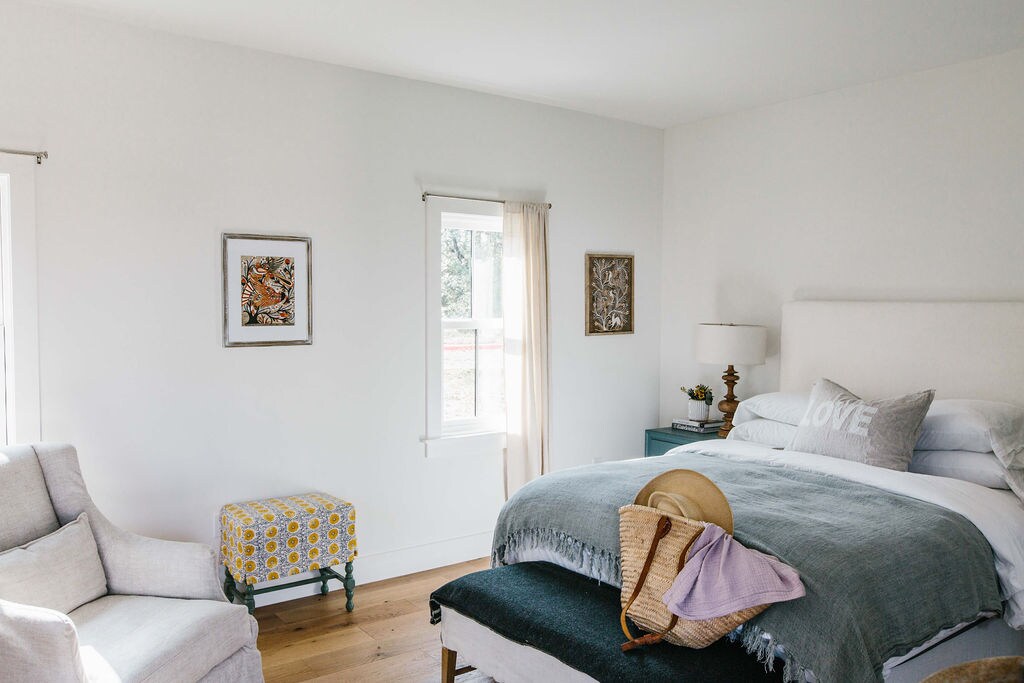A bright and airy bedroom features a spacious bed adorned with a soft throw and decorative pillows. Natural light streams in through two windows, illuminating subtle wall art. A cozy armchair and a colorful stool provide additional seating, enhancing the room's relaxed ambiance.