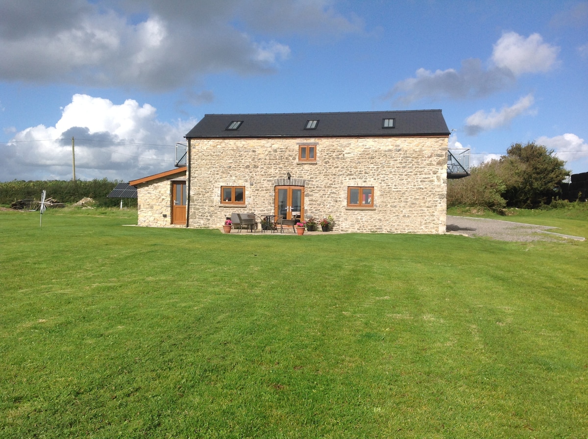 A rustic stone cottage is pictured, surrounded by a green paddock. The building features large windows and a mix of contemporary and traditional architectural elements. A small outdoor seating area with potted plants is visible in front, set against a backdrop of partly cloudy skies.