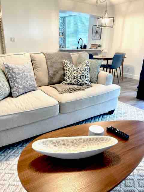 A light-colored, comfortable couch is positioned in a cozy living area, adorned with decorative pillows. A wooden coffee table holds a decorative bowl and a remote control. In the background, dining chairs and a table are visible, contributing to the unit's inviting layout.