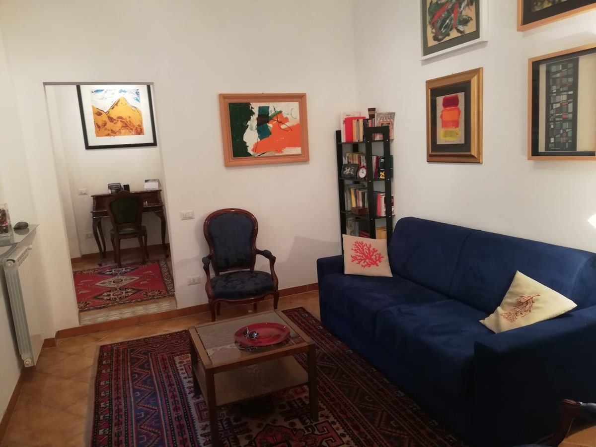 A cozy living area is shown, featuring a blue sofa adorned with decorative pillows. A wooden coffee table sits in front of the sofa, surrounded by a patterned rug. A reading chair and shelves filled with books enhance the inviting space. Artwork adorns the walls.