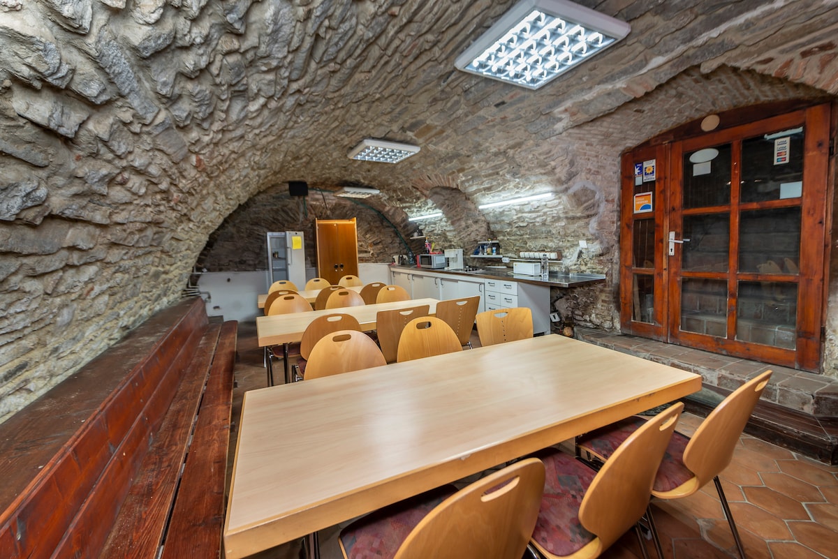 An arched stone ceiling creates a unique ambiance in the communal dining area. A long table is surrounded by wooden chairs, and kitchen appliances are arranged on counters against the wall. Natural light filters through illuminated panels above.