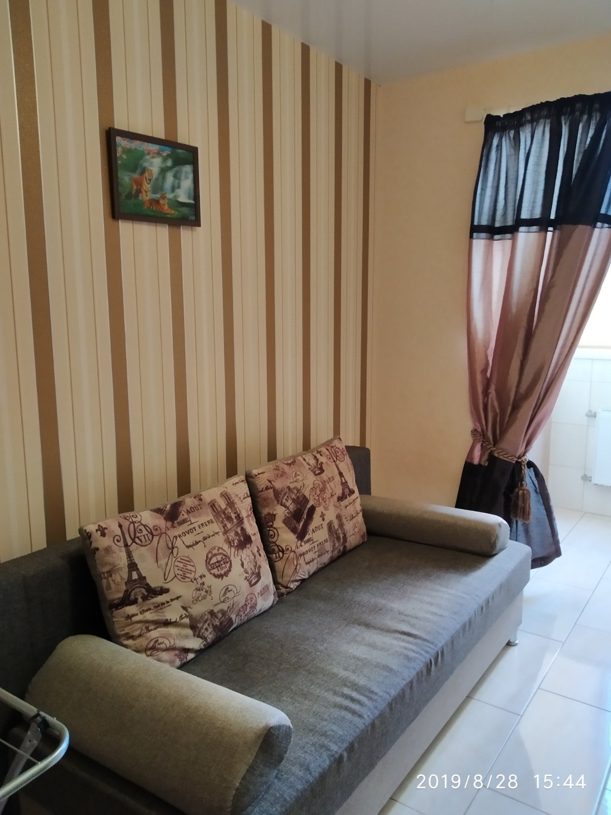 A seating area features a gray sofa adorned with decorative cushions. The walls are lined with vertical beige and brown stripes. A small framed artwork hangs above the sofa, and a curtain with a rich color contrasts against the light walls.