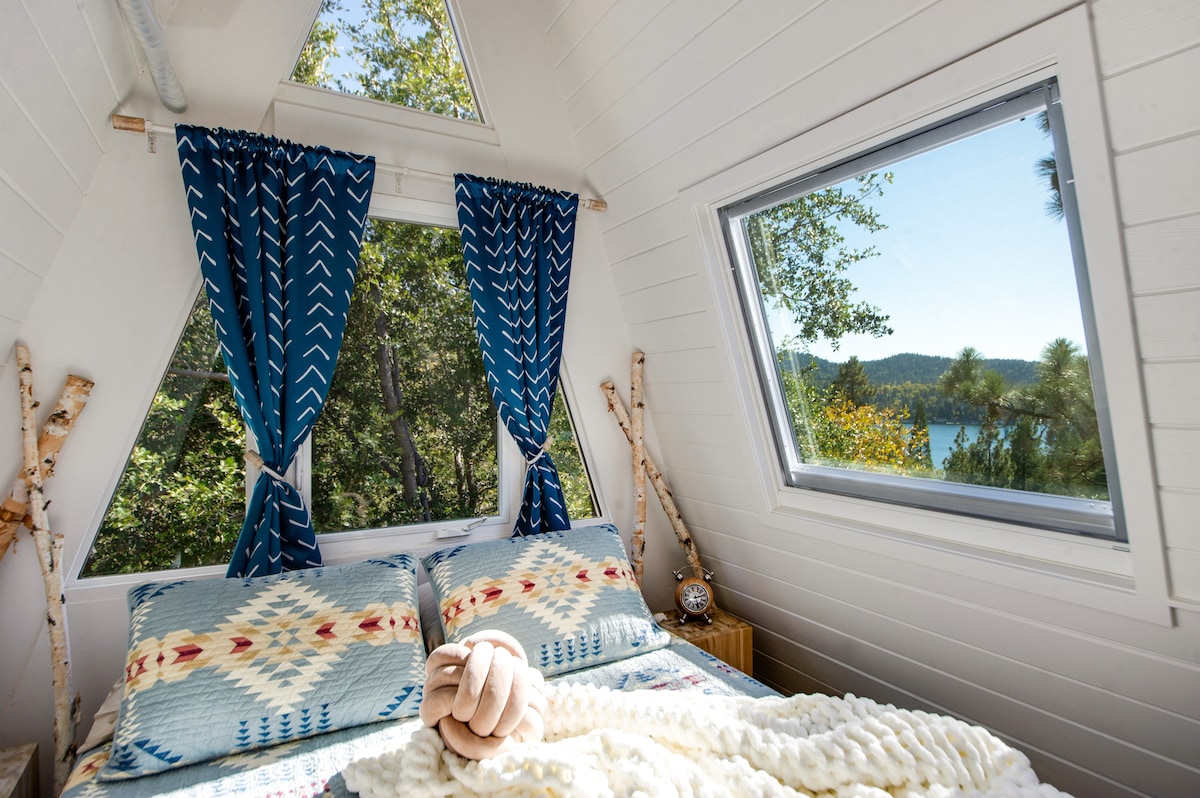A comfortable sleeping area is featured with a cozy bed adorned with patterned throws. Large windows provide scenic views of the surrounding trees and lake. Soft blue curtains frame the windows, allowing natural light to fill the room.
