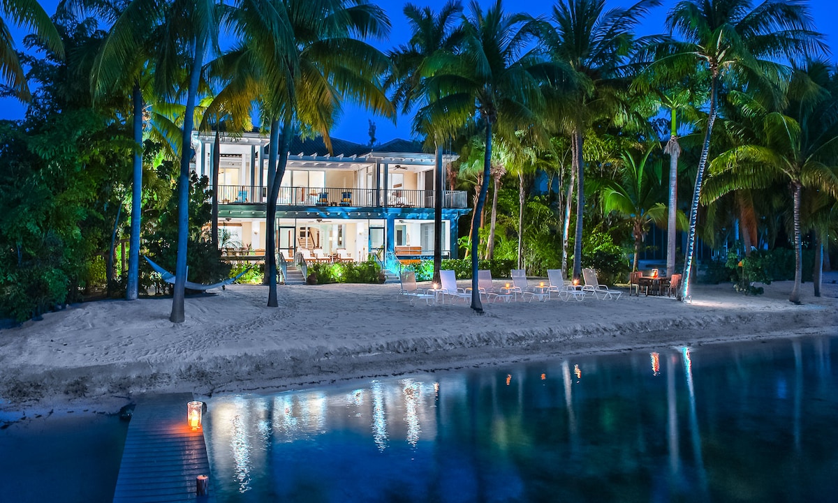 The beachfront villa is illuminated at night, revealing a serene exterior framed by palm trees. Soft lights reflect off the water, enhancing the tranquil ambiance. Outdoor seating is visible on the sand, inviting relaxation under the starry sky.