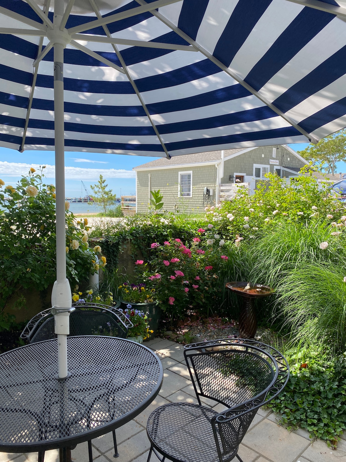 A private courtyard is showcased with a round table and two black metal chairs beneath a striped umbrella. Lush greenery and colorful flowers surround the area, offering a serene setting. In the background, a glimpse of the ocean can be seen, enhancing the tranquil atmosphere.