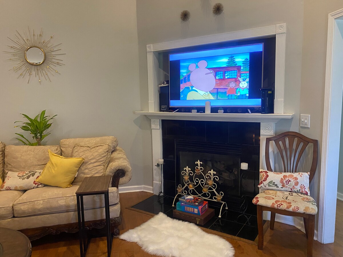 A cozy living room features a soft couch and decorative pillows positioned near a fireplace. A small table stands beside the couch, and a decorative plant adds a touch of greenery. A television is mounted above the fireplace, displaying a cartoon.