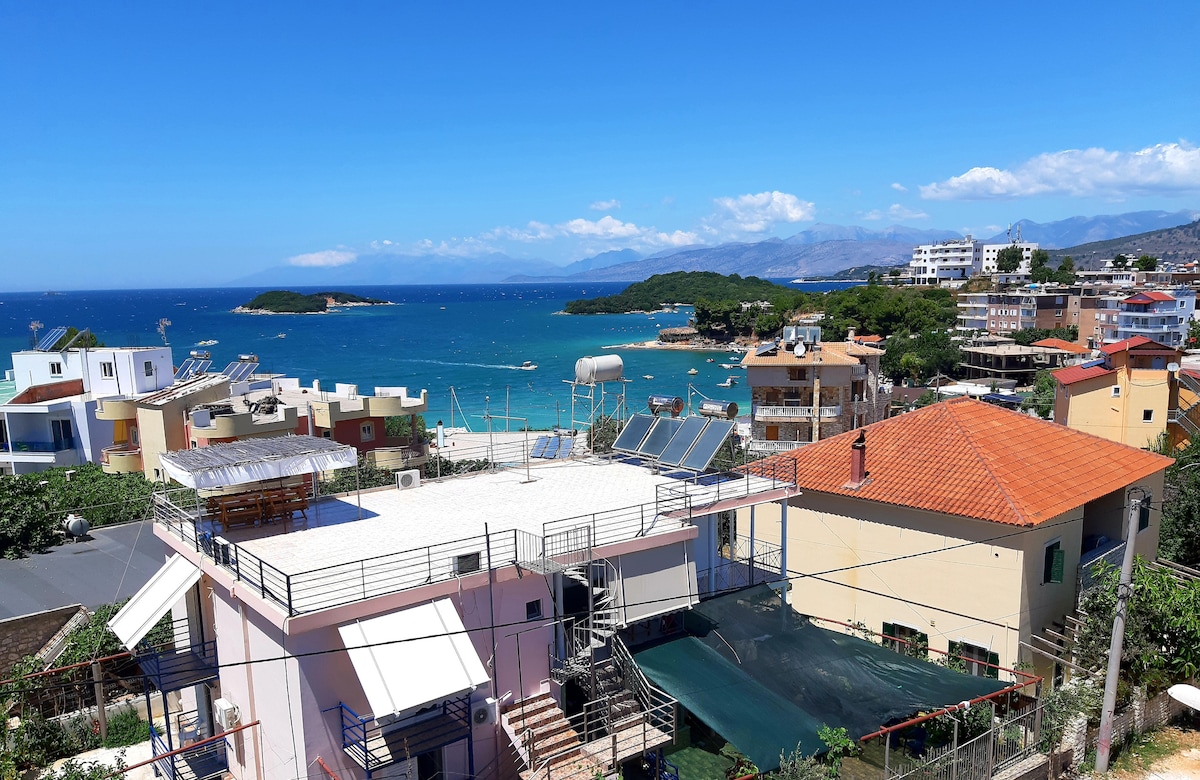 A panoramic view displays the coastline and azure waters of Ksamil. The rooftops of neighboring buildings are visible, with the vibrant blue sea and distant islands enriching the scene. Clear skies provide a bright backdrop for this tranquil coastal setting.