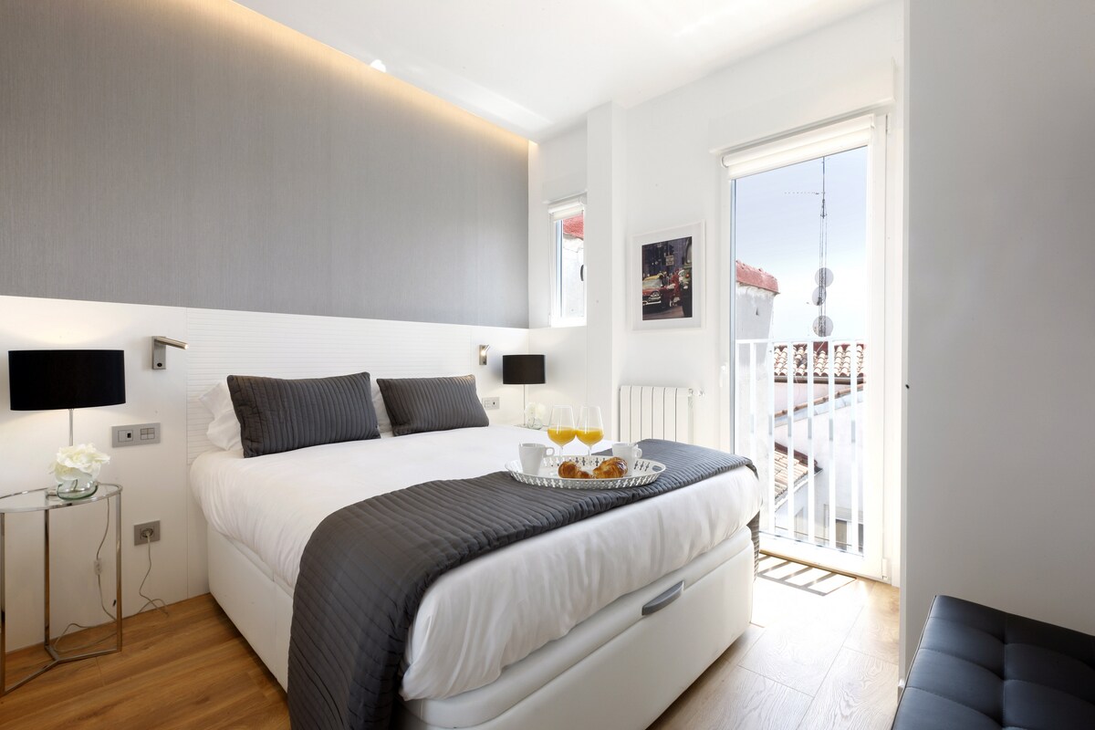 A bright bedroom features a spacious bed with a neatly arranged gray blanket. A small table holds a tray with breakfast items. Natural light enters through a glass door leading to a small balcony, which is adorned with white railings.