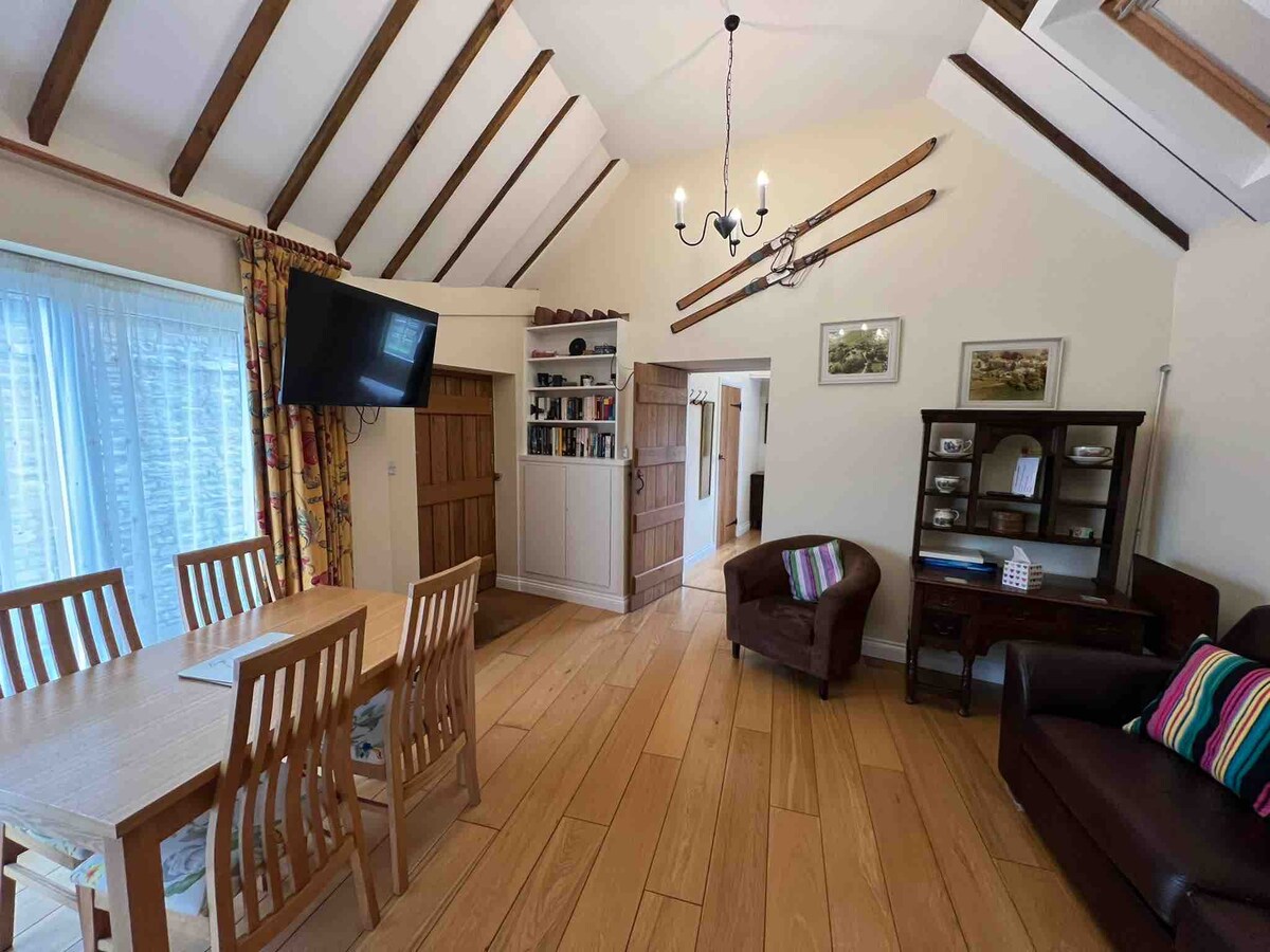 An open living space is highlighted by exposed wooden beams and large windows allowing natural light. A dining table with four chairs is positioned near the window. A cozy brown sofa and a tub armchair are arranged to create a welcoming seating area.