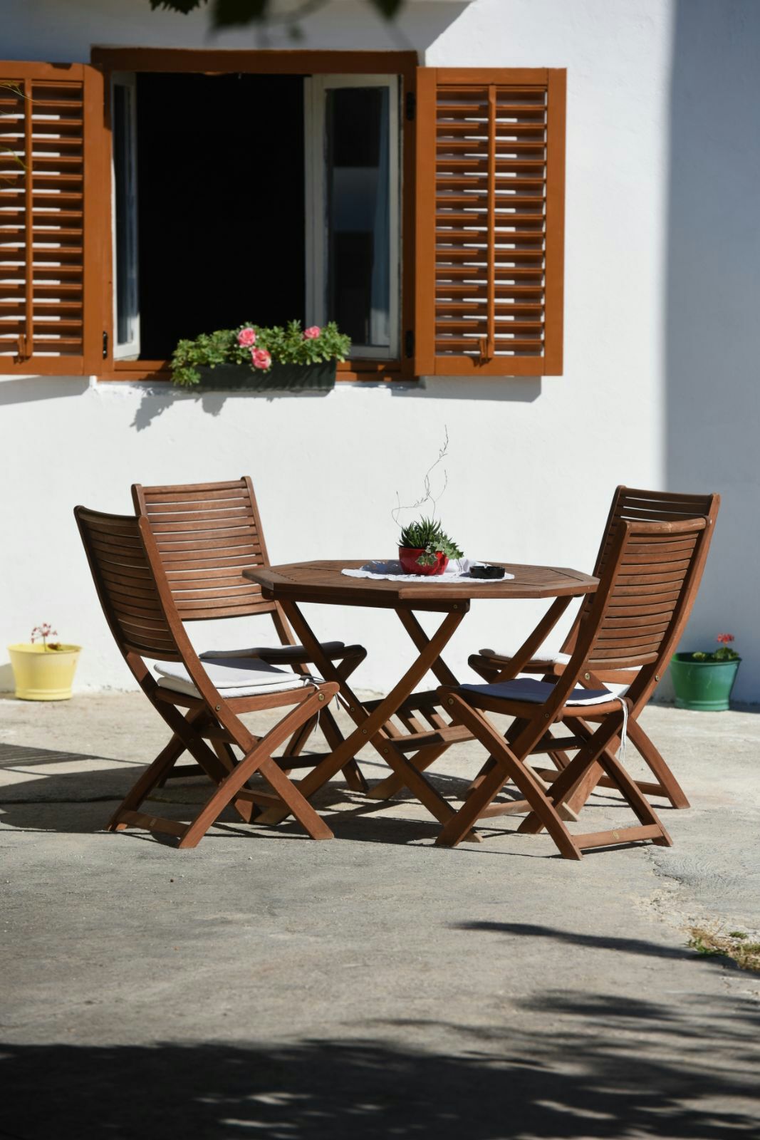 A wooden table surrounded by three folding chairs is positioned in an outdoor area. Sunlight illuminates the space, highlighting the brown tones of the furniture. Shuttered windows frame the scene, with a small plant placed at the center of the table, adding a touch of greenery.