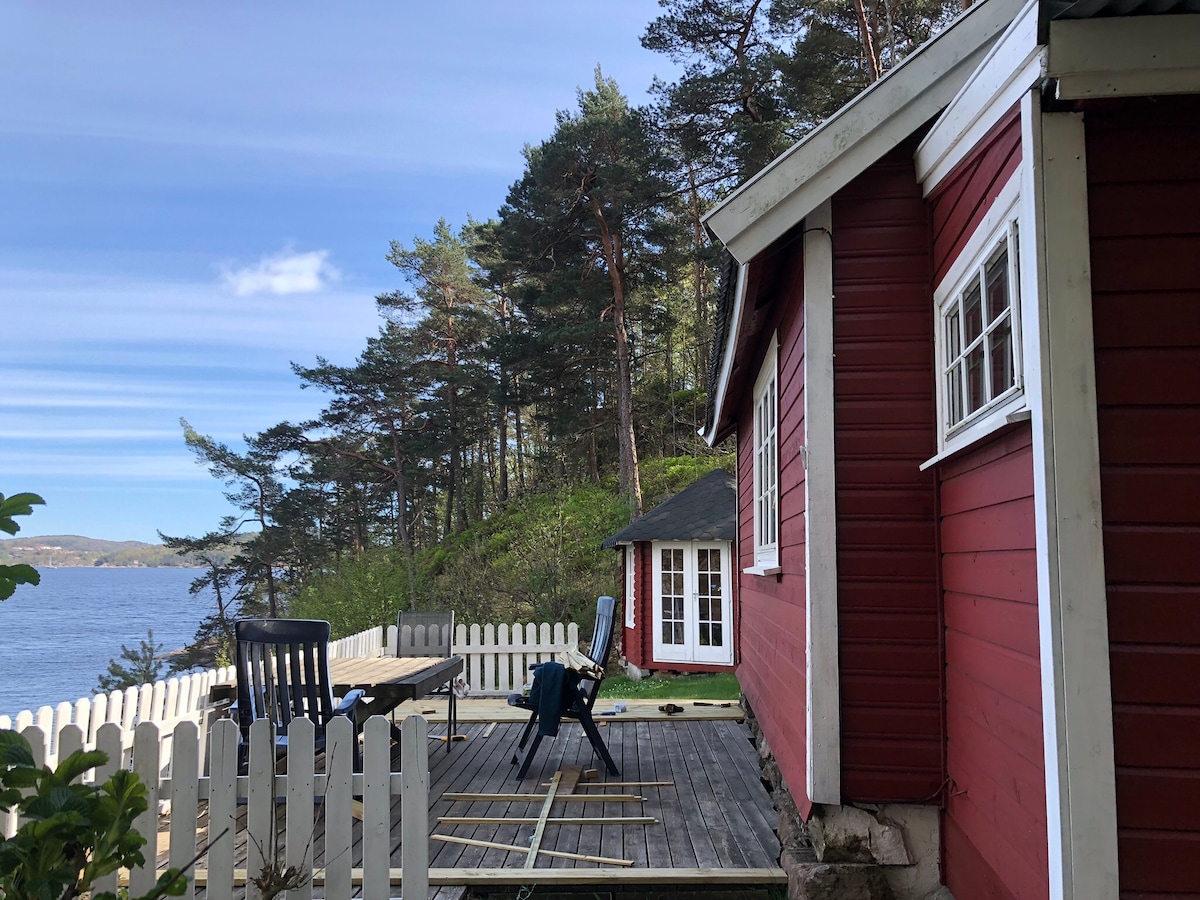The exterior of a red cabin is displayed, showcasing a spacious deck with seating. Surrounding trees provide a natural backdrop, while a glimpse of the nearby water is visible. The cabin features distinctive windows, adding charm to the rustic architecture.