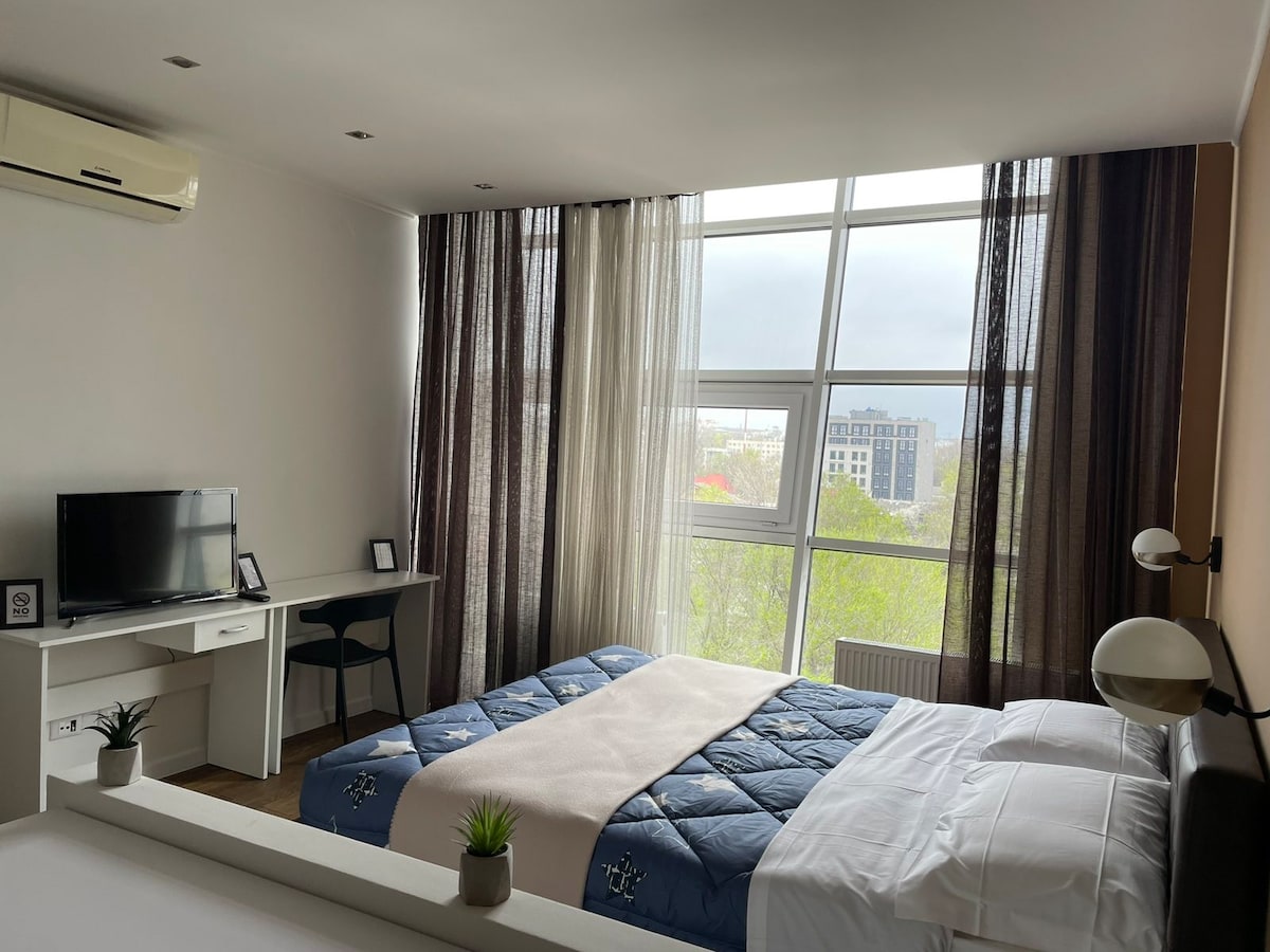 A sleeping area features a spacious bed with a blue quilt and white linens. Natural light floods through large windows covered by sheer curtains and dark drapes. A compact desk and chair are positioned nearby, with a television mounted on the wall, enhancing the functionality of the space.