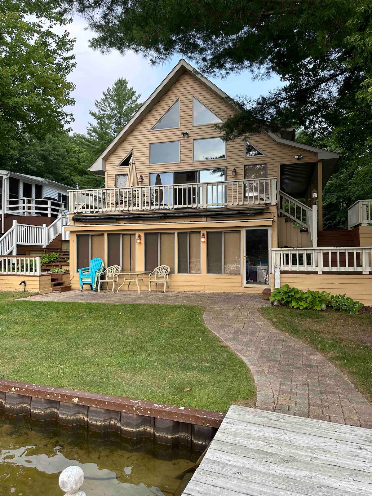 The exterior of the house is showcased with a triangular roof and multiple large windows. A spacious deck extends from the front, featuring chairs and a table. A brick pathway leads to the lawn, which borders the clear water of the lake.