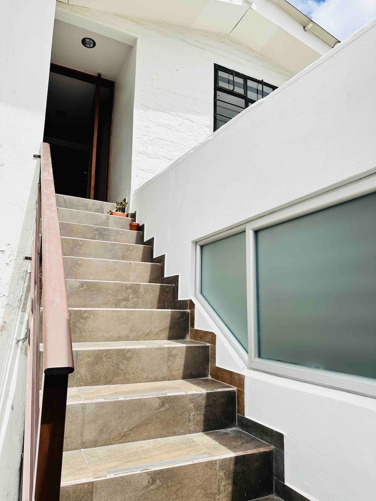 A well-lit staircase leads to the entrance, featuring tiled steps with a mix of neutral tones. A handrail runs alongside for support, while a sleek window allows natural light to brighten the space, creating an inviting entryway.