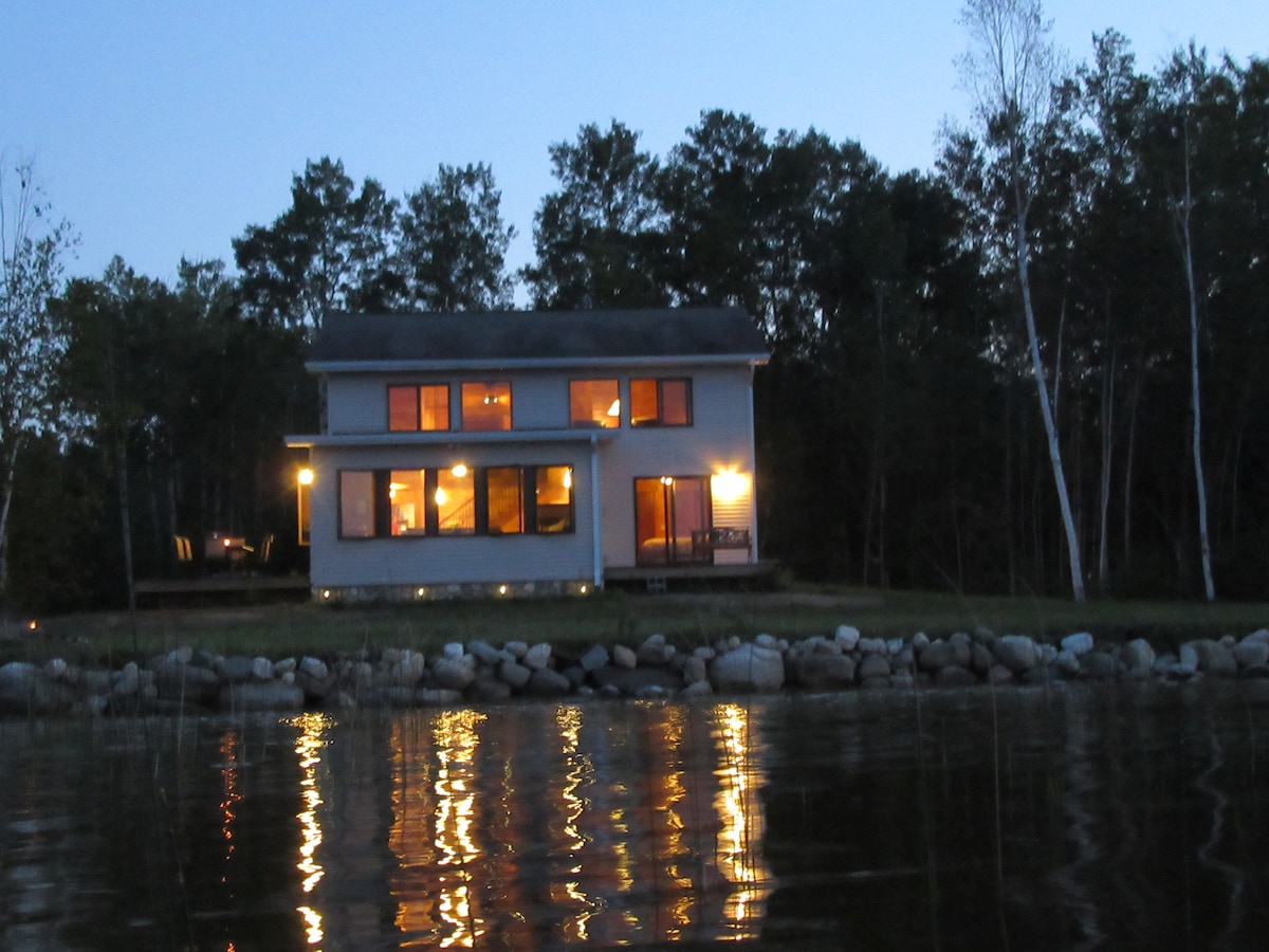 The exterior of the lake house is illuminated at dusk, showcasing large windows that reflect warm light against the water. A stone shoreline borders the property, with surrounding trees creating a natural backdrop, enhancing the serene atmosphere of the location.