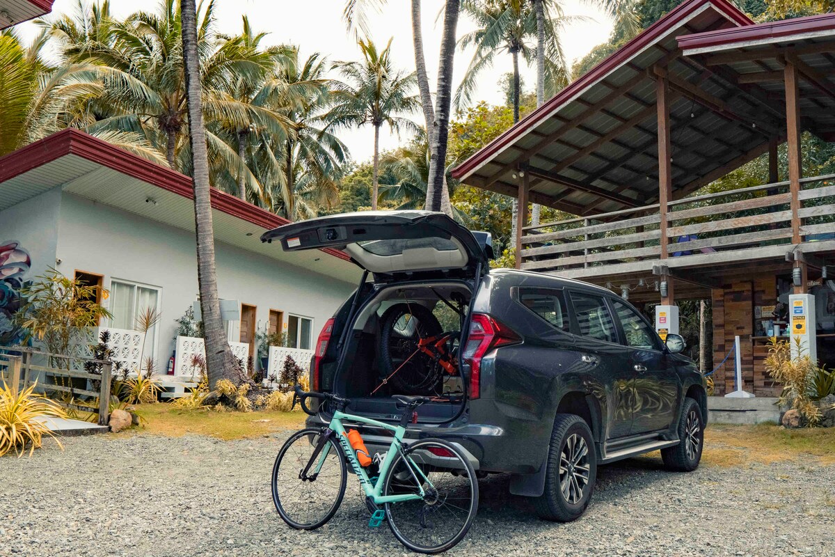 A vehicle is parked with its rear open, displaying a bicycle inside. Palm trees are visible in the background, alongside a two-story building featuring a balcony. The exterior is surrounded by gravel and tropical vegetation, creating a blend of outdoor and accommodation elements.