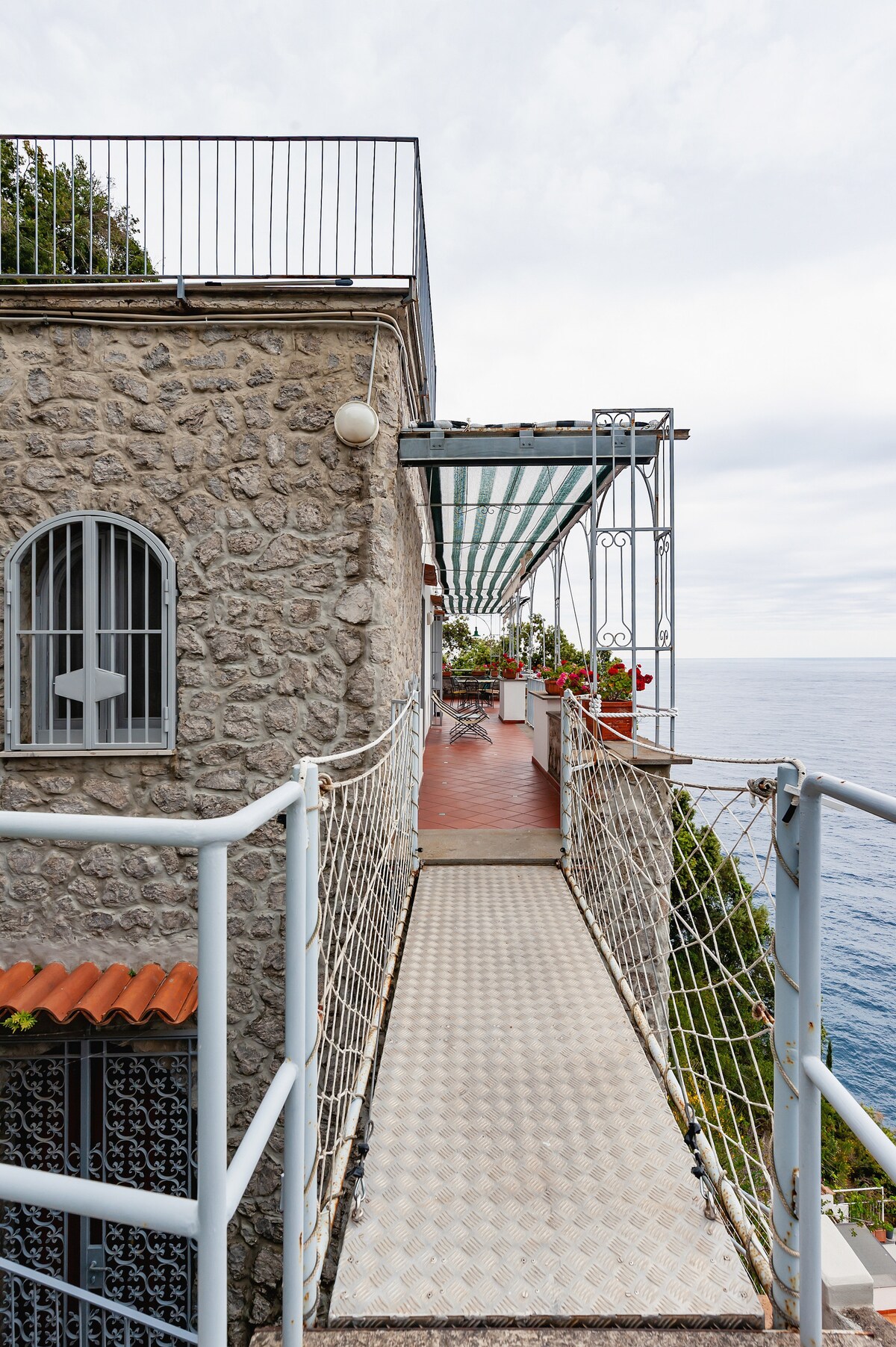 A narrow walkway with a metal grating surface extends along the exterior of the villa. It is flanked by stone walls and features decorative potted plants. The pathway leads toward an area with a retractable awning, providing views of the sea beyond.