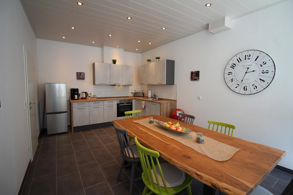 A modern kitchen space features light wood cabinetry and gray accents. A large wooden dining table with colorful chairs accommodates guests. Stainless steel appliances are positioned against the wall, complemented by ample countertop space. Soft lighting highlights the open layout.