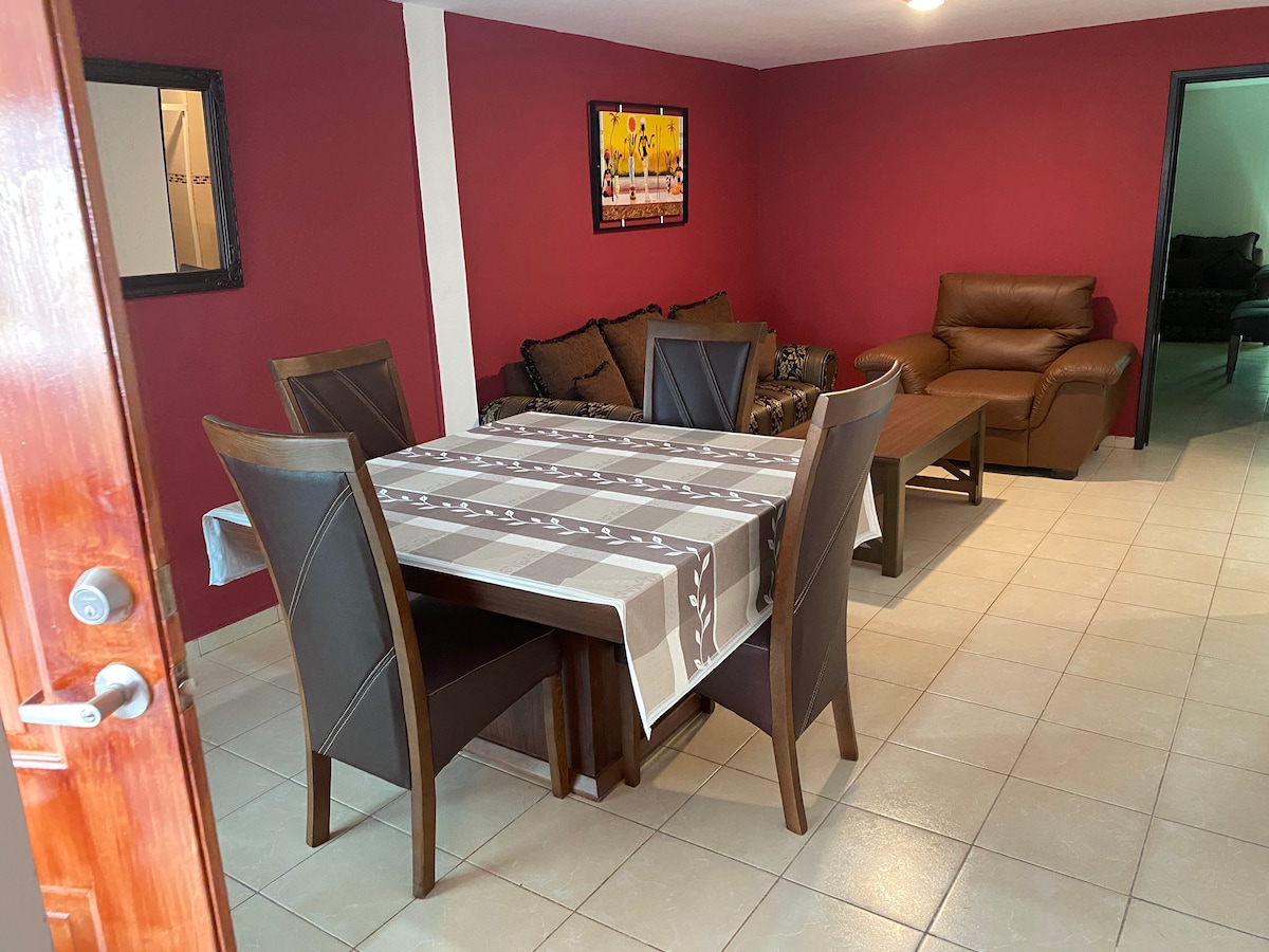 A dining area is depicted, featuring a large table surrounded by six dark chairs. The walls are painted in a rich burgundy color. A comfortable loveseat and an armchair can be seen in the background, contributing to a relaxed setting. A piece of artwork is displayed on the wall.