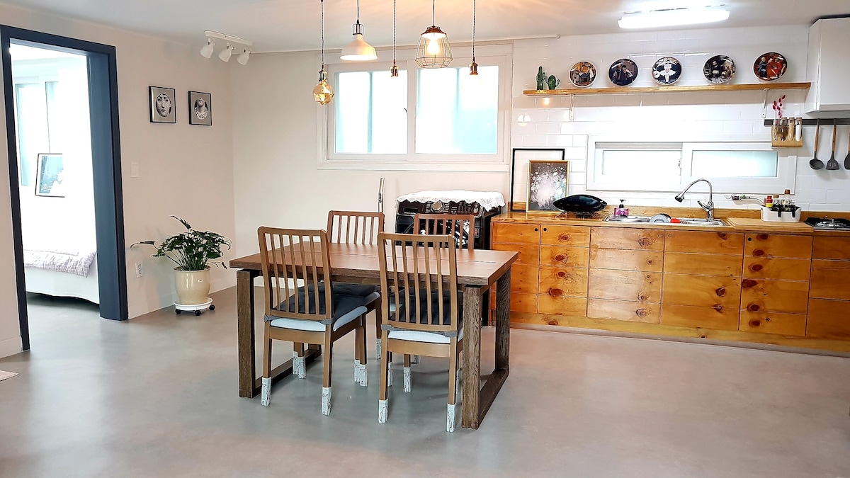A spacious kitchen and dining area features a wooden table with four chairs, positioned on a light concrete floor. Wall shelves display decorative items, while ample natural light floods in through large windows. Minimalist cabinetry and modern appliances create a functional cooking space.