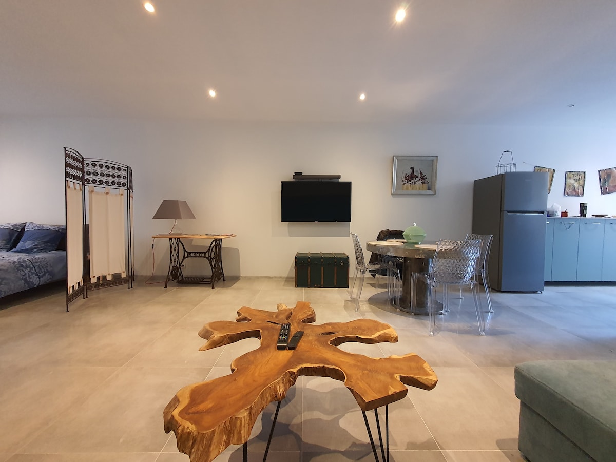 An open living space features a unique wooden coffee table at its center, surrounded by a spacious sofa and a dining area. A modern television is mounted on the wall, with a refrigerator and kitchen area visible in the background. Natural light brightens the neutral-toned walls and flooring.