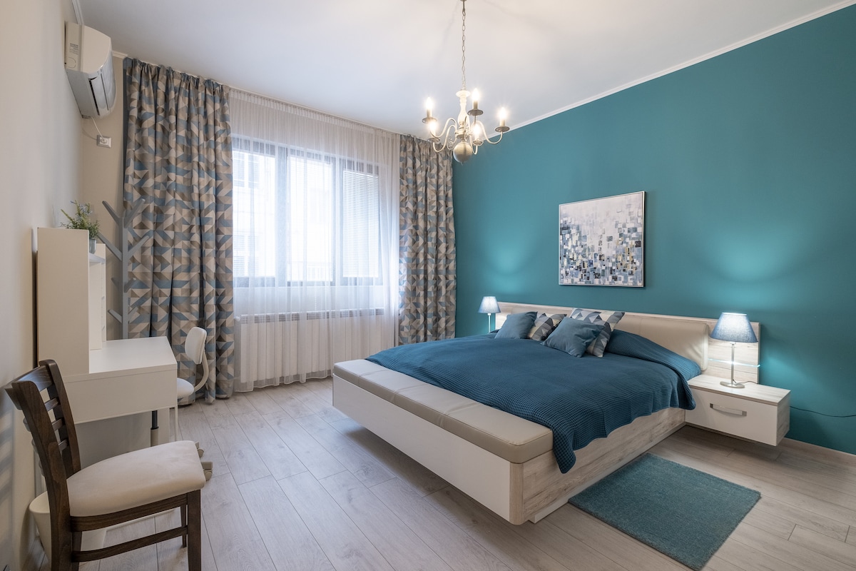 A bright and spacious bedroom features a large, comfortable bed with a blue blanket. Two bedside lamps provide soft lighting, while a piece of abstract art adorns the wall. A desk and chair are positioned by a window dressed in sheer curtains, allowing natural light to fill the room.