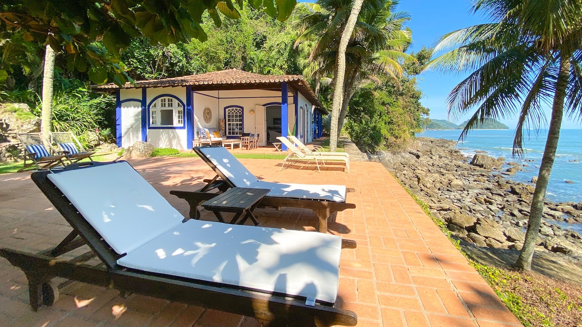 A serene outdoor space features lounge chairs positioned near the rocky shoreline. The house, with its blue and white facade, is set amidst lush greenery and palm trees. The calm sea stretches across the background under a clear blue sky.