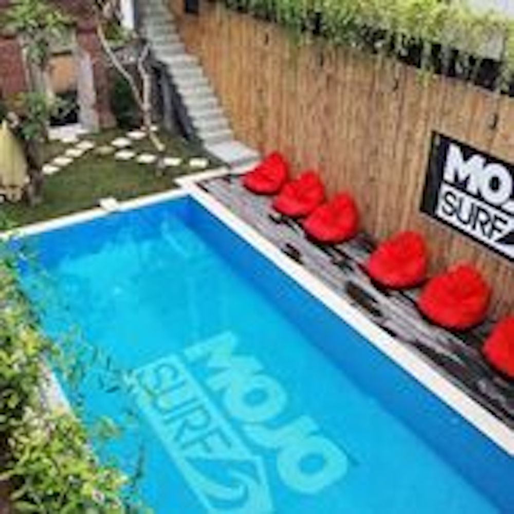 A swimming pool reflects the surroundings, featuring a logo displayed on the bottom. Colorful bean bags are arranged along the edge, offering relaxation spaces. Lush greenery and a walkway lead to a serene outdoor area, enhancing the space's inviting atmosphere.
