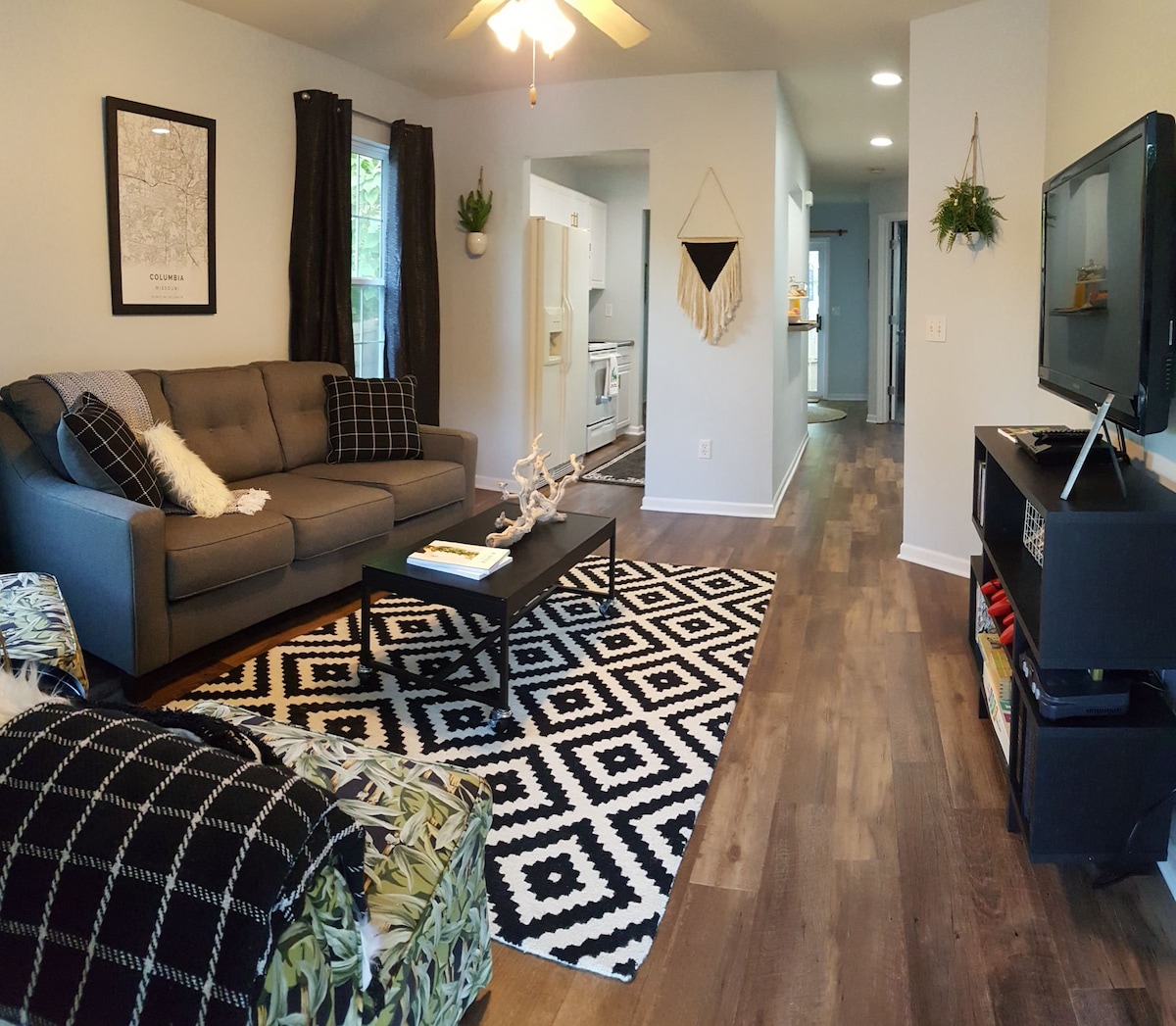The living room features a modern sofa and patterned armchair, complemented by a geometric area rug. A coffee table sits in front, with a flat-screen TV on a nearby shelf. Natural light enters through windows dressed with dark curtains, enhancing the inviting ambiance.