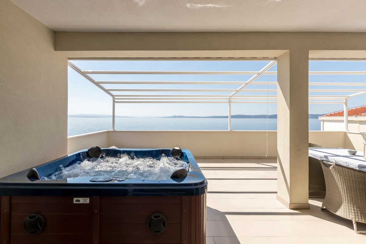 A spacious terrace features a hot tub with a view of the sea, surrounded by light-colored walls and a canopy providing shade. Nearby, a seating area with wicker chairs offers a comfortable space to relax while enjoying the serene ocean scenery.