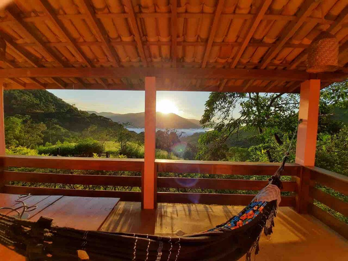 A spacious porch features a hammock and wooden railing, overlooking a landscape bathed in warm sunlight. The view includes rolling hills and distant mountains, creating a serene atmosphere. A thatched roof complements the natural setting, enhancing the connection with the outdoors.