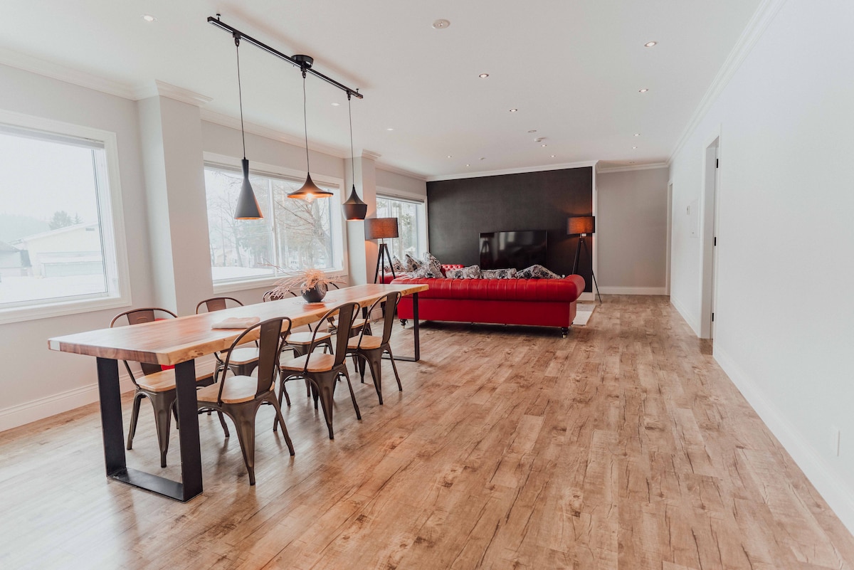 A large dining area features a long wooden table surrounded by eight chairs, complemented by stylish pendant lighting above. Opposite the table, a red sofa is positioned against a dark accent wall, with large windows providing natural light and views of the surrounding area.