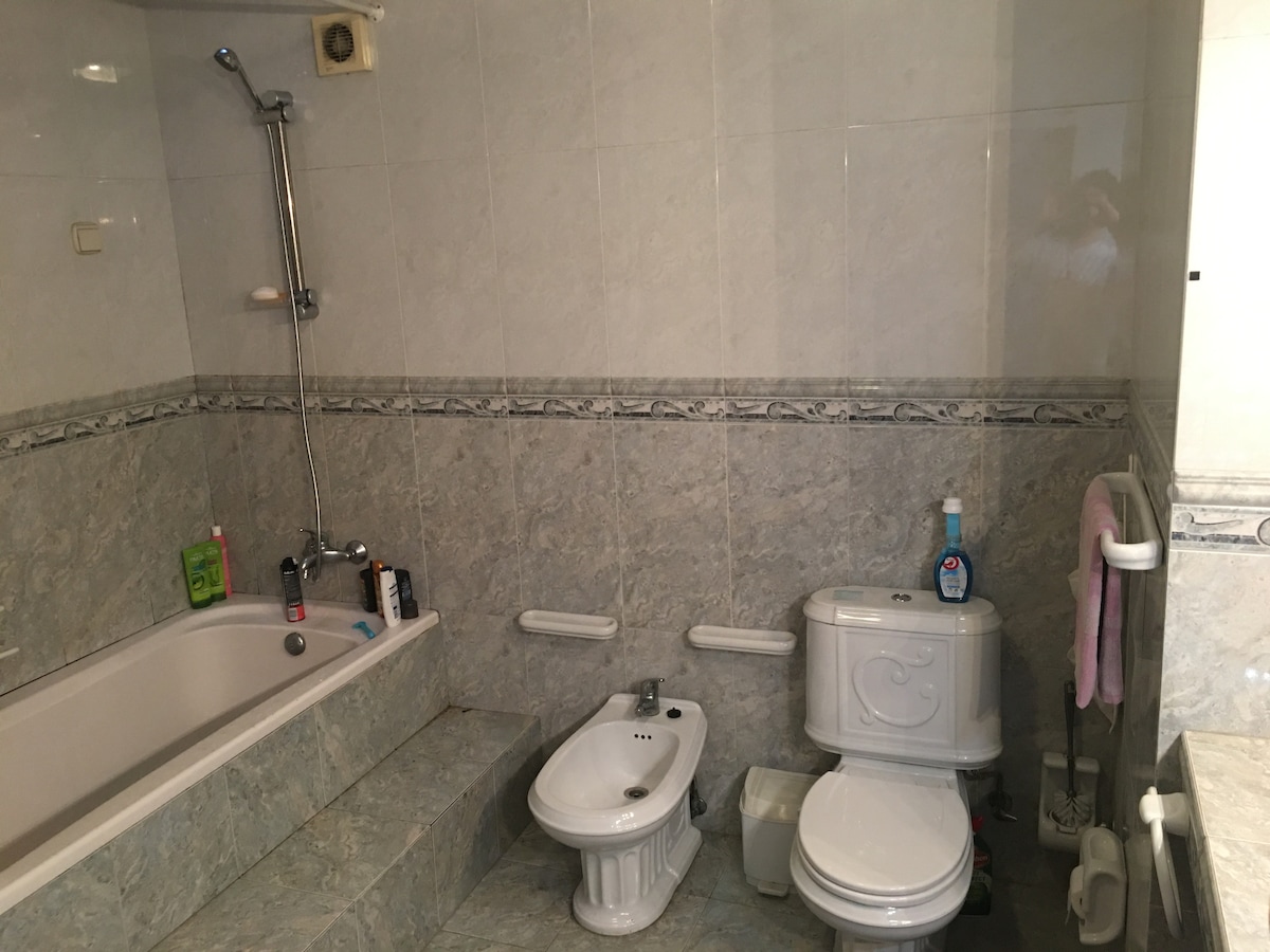 The shared bathroom features a bathtub with an attached shower, a bidet, and a toilet, all set against marble-effect tiles. Shelving along the wall holds toiletries, and grab bars are installed near the bathtub for accessibility.